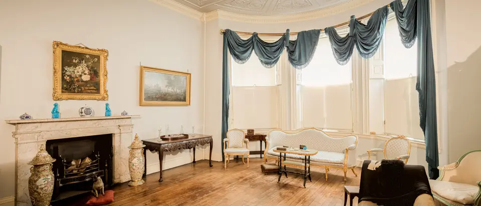 A bright, old-fashioned living room with a marble fireplace and tall curtained windows