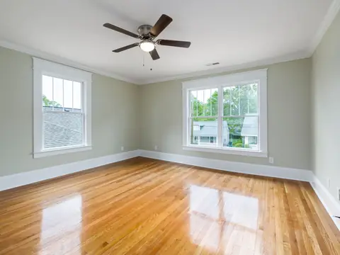 An empty room with a polished wood floor, a ceiling fan and two bright windows