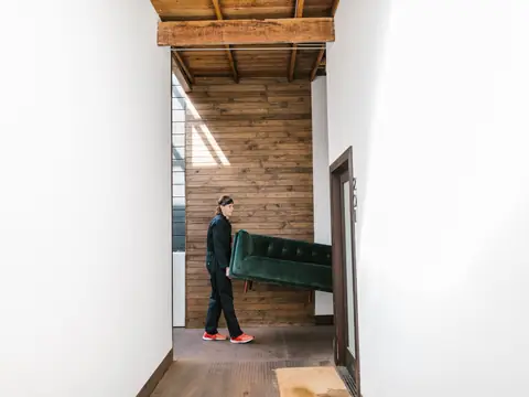 A man carrying a green sofa down a bright hallway