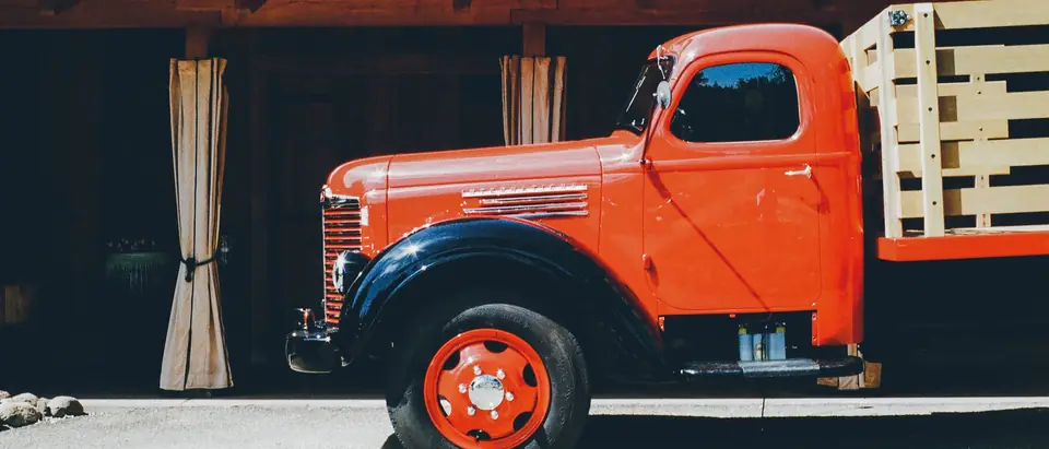 An orange vintage truck with a wooden stake bed parked in the sun