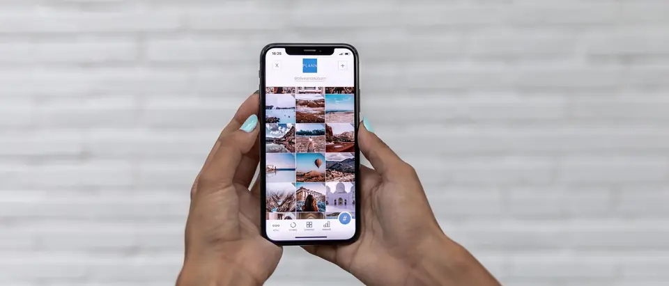 Two hands holding a smartphone showing a grid of photos against a white brick wall