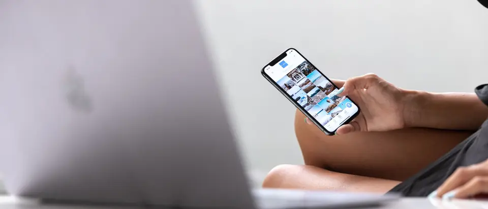 A hand holding a smartphone beside an open laptop on a white desk