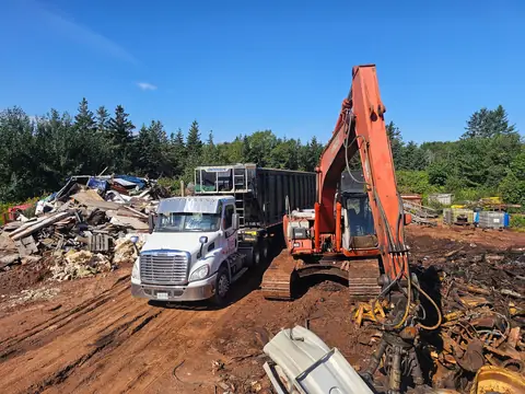 A white truck and an orange excavator at a scrap yard beside piles of cleared metal and debris