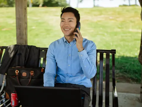 A man on a phone call on a park bench with a laptop on a sunny day