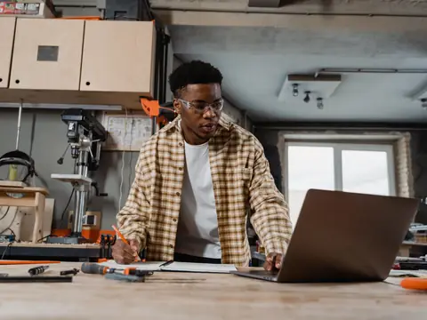 A man working on a laptop at a bench in a bright workshop